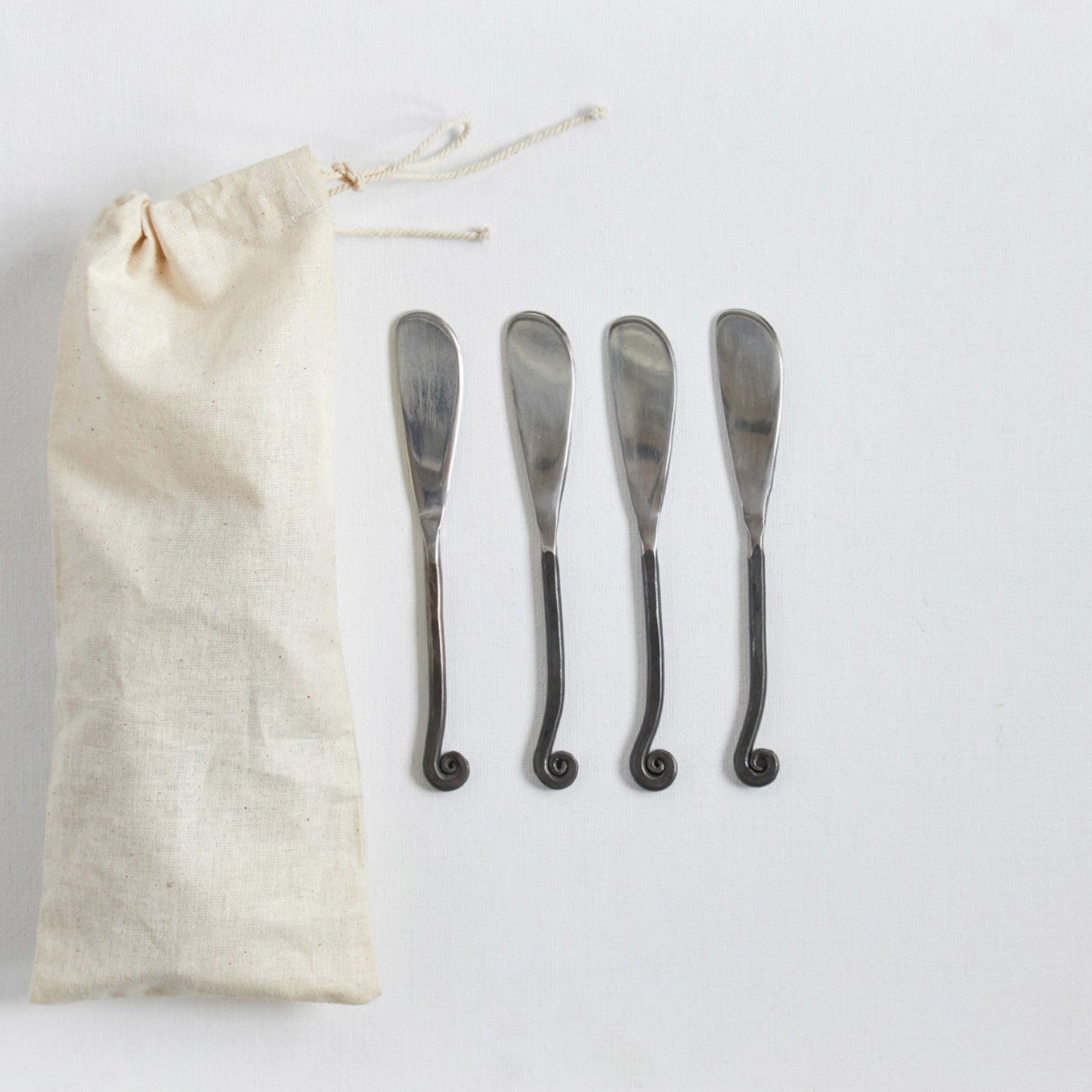 A set of four hand-forged silver canape knives displayed alongside a beige fabric pouch on a white background.